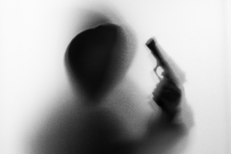 Shadow blur of horror man in jacket with hood gun in his hand.Dangerous man behind the frosted glass.Mystery man.Halloween background.Black and white picture.Blur pictureの写真素材