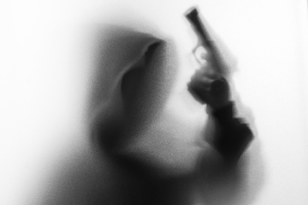 Shadow blur of horror man in jacket with hood gun in his hand.Dangerous man behind the frosted glass.Mystery man.Halloween background.Black and white picture.Blur pictureの写真素材