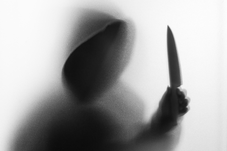 Shadow blur of horror man in jacket with hood knife in his hand.Dangerous man behind the frosted glass.Mystery man.Halloween background.Black and white picture.Blur pictureの写真素材