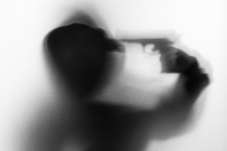 Shadow blur of horror man in jacket with hood gun in his hand.Dangerous man behind the frosted glass.Mystery man.Halloween background.Black and white picture.Blur pictureの写真素材