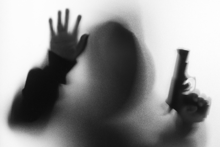 Shadow blur of horror man in jacket with hood gun in his hand.Dangerous man behind the frosted glass.Mystery man.Halloween background.Black and white picture.Blur pictureの写真素材