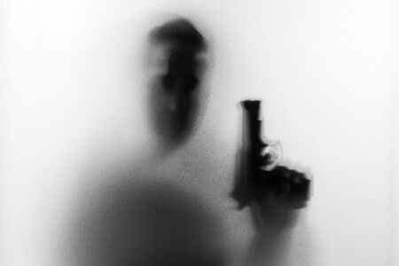 Shadow of horror man killer with a gun in his hand.Dangerous man behind the frosted glass.Mystery man.Halloween background.Black and white picture.Blur pictureの写真素材