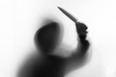 Shadow blur of horror man in jacket with hood knife in his hand.Dangerous man behind the frosted glass.Mystery man.Halloween background.Black and white picture.Blur pictureの写真素材