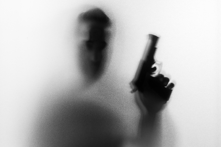 Shadow of horror man killer with a gun in his hand.Dangerous man behind the frosted glass.Mystery man.Halloween background.Black and white picture.Blur pictureの写真素材