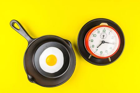 Frying pan with fried egg and alarm clock in a black plate isolated on a yellow background viewed from above.Concept of breakfast at the race against time.の写真素材