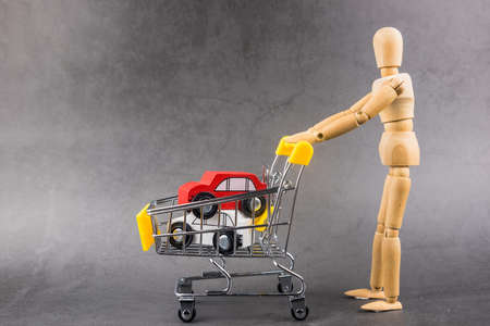 A wooden doll carries a red car model in metal shopping cart. Man buy his new car in dealership....
