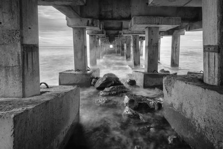 Black and white picture of bottom view of a rocky coast boarding an old pier during a dramatic sunset.の写真素材