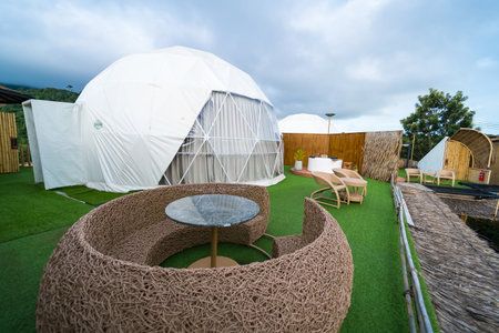 a modern geodesic dome tent situated on a lush green lawn, under a vast blue sky punctuated by fluffy white clouds. The domeâs white fabric cover reflects the sunlight.の写真素材