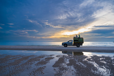 A garbage collection truck is seen on a beach with two sanitation workers sitting on top of the waste container. The scene highlights the efforts to maintain cleanliness and manage waste in beach.のeditorial素材
