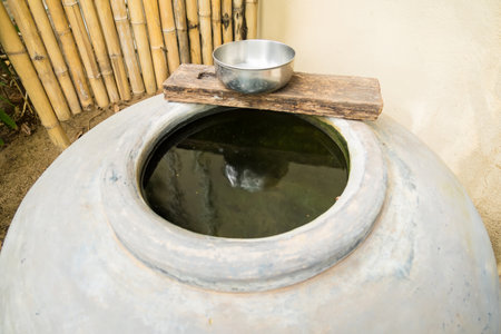 A traditional ceramic water storage jar with a rustic wooden lid and a metal bowl on top, set...