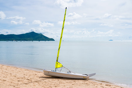 A yellow sailboat on a sandy beach with calm waters and distant mountains under a partly cloudy sky.の写真素材