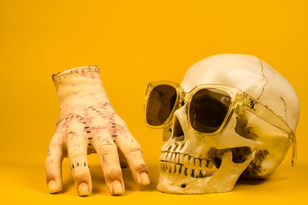 A human skull and severed hand with sunglasses on a bright yellow backdrop, adding a spooky touch to Halloween decor.の写真素材