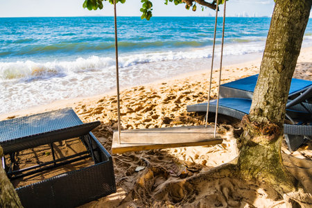A serene beach scene featuring a wooden swing hanging from a tree, overlooking the ocean waves with relaxing loungers nearby.の写真素材