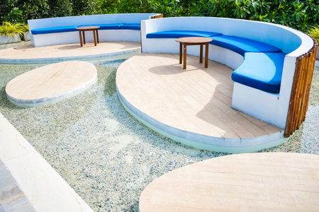 A contemporary outdoor seating area with curved wooden platforms, blue cushioned benches, and small wooden tables, surrounded by lush greenery for a relaxing ambiance.の写真素材