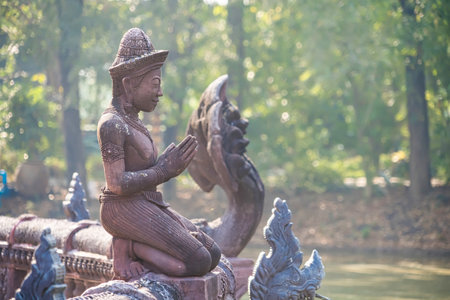 Lop Buri, Thailand - January, 02, 2025 : A carved statue of a praying figure on a bridge with ornate dragon sculptures in a sunlit forest setting at Lop Buri, Thailand.の写真素材