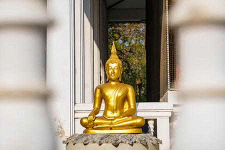 Golden Buddha statue in a serene meditative pose at a temple surrounded by sunlight and architectural featuresの写真素材