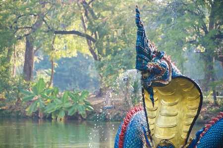 Lop Buri, Thailand - January, 02, 2025 : Ornate giant serpent statue in a serene park pond surrounded by lush green trees on a sunny day at Lop Buri, Thailand.の写真素材