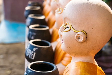 Lop Buri, Thailand - January, 02, 2025 :Colorful ceramic monk statues in orange robes holding alms bowls displayed on a stone surface in bright daylight at Lop Buri, Thailandの写真素材