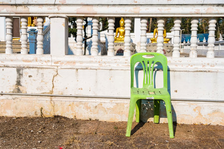 A lone green plastic chair placed in front of a white balustrade with golden Buddha statues on a sunny dayの写真素材