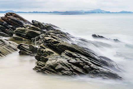 Photo long exposure of Rocky shoreline with jagged rocks extending into the calm sea under a cloudy sky, creating a serene coastal landscapeの写真素材