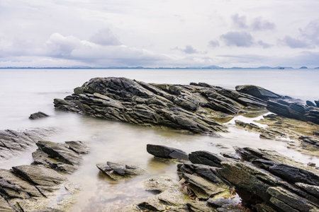 Photo long exposure of Rocky shoreline with jagged rocks extending into the calm sea under a cloudy...