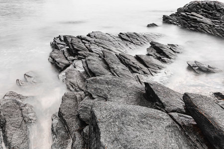 Black and white photo long exposure of Black rugged rocks extend into the misty waters, creating a dramatic coastal landscape with overcast skiesの写真素材