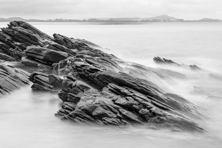 Black and white photo long exposure of Black rugged rocks extend into the misty waters, creating a dramatic coastal landscape with overcast skiesの写真素材