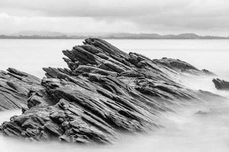 Black and white photo long exposure of Black rugged rocks extend into the misty waters, creating a dramatic coastal landscape with overcast skiesの写真素材