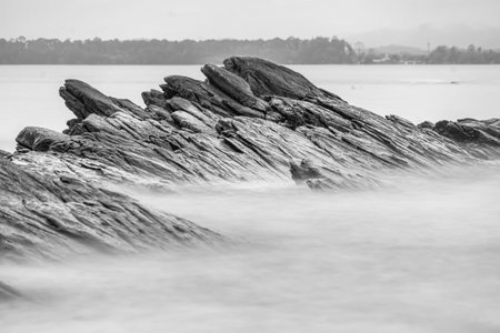 Black and white photo long exposure of Black rugged rocks extend into the misty waters, creating a dramatic coastal landscape with overcast skiesの写真素材