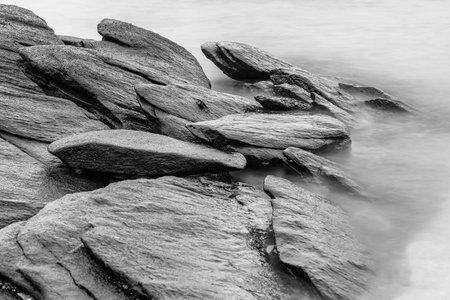 Black and white photo long exposure of rugged rocks extend into the misty waters, creating a dramatic coastal landscape with overcast skiesの写真素材