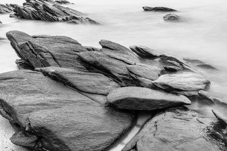 Black and white photo long exposure of Black rugged rocks extend into the misty waters, creating a dramatic coastal landscape with overcast skiesの写真素材