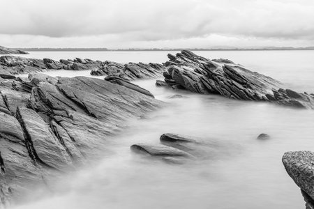 Black and white photo long exposure of Black rugged rocks extend into the misty waters, creating a dramatic coastal landscape with overcast skiesの写真素材