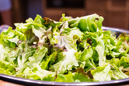A close-up view of a large metal bowl filled with fresh, mixed green lettuce leaves in a kitchen settingの写真素材