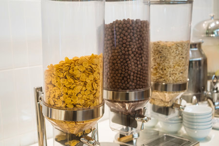 Close-up view of cereal dispensers filled with cornflakes, chocolate balls, and oats in a breakfast buffet settingの写真素材