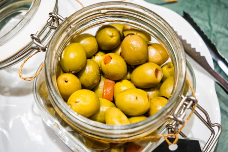 Close-up of a glass jar filled with fresh green olives and a small red pepper piece, placed on a white plate with cutlery nearbyの写真素材