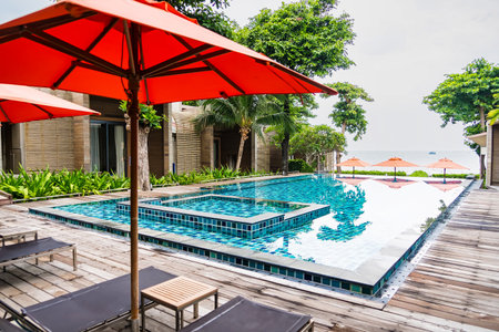 Rayong, Thailand - April, 03, 2025 :Luxurious outdoor swimming pool with red umbrellas and sun loungers at a tropical resort, surrounded by lush greeneryの写真素材