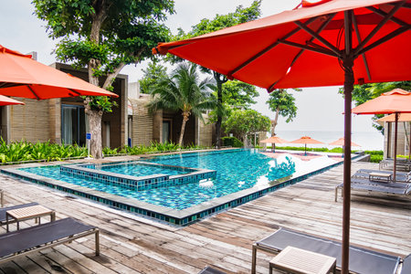 Rayong, Thailand - April, 03, 2025 :Luxurious outdoor swimming pool with red umbrellas and sun loungers at a tropical resort, surrounded by lush greeneryの写真素材