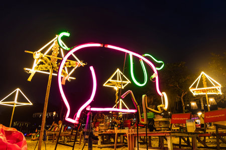 Rayong, Thailand - April, 01, 2025 :Neon pig sign and decorative illuminated umbrellas on a beach at night, with people sitting on red bean bags in backgroundの写真素材