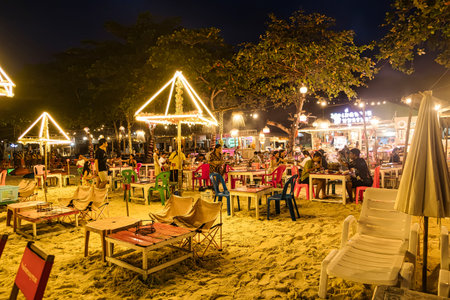 Rayong, Thailand - April, 01, 2025 : Night beach scene with illuminated tables and chairs, people enjoying a lively atmosphere and tropical environment.の写真素材