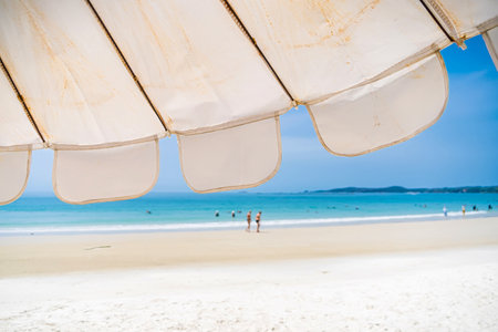 Rayong, Thailand - April, 01, 2025 : Beach view under a large umbrella with distant walkers along the shore on a sunny day at an idyllic coastal locationの写真素材
