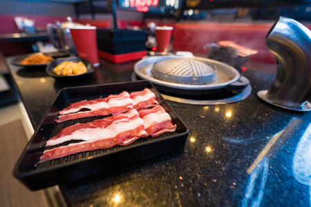 Inside a Korean barbecue restaurant, raw bacon strips are ready to be grilled on a tabletop stove at a dining tableの写真素材