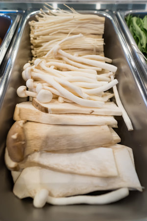 Various fresh mushrooms including enoki and king oyster neatly arranged in a stainless steel tray at...