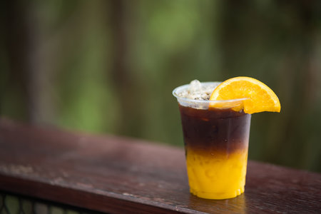 Plastic cup of iced coffee with orange juice and an orange slice on a wooden railing against a blurred natural backgroundの写真素材