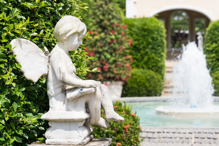 A stone angel sculpture with wings sits near a lush hedge beside a bubbling fountain in a tranquil garden settingの写真素材