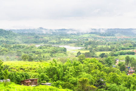 Lush green valley landscape with misty hills, dense trees, and scattered houses under a cloudy skyの写真素材