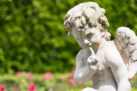 A stone angel sculpture with wings sits near a lush hedge beside a bubbling fountain in a tranquil garden settingの写真素材