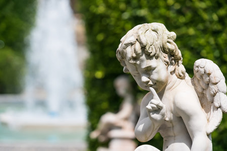 A stone angel sculpture with wings sits near a lush hedge beside a bubbling fountain in a tranquil garden settingの写真素材