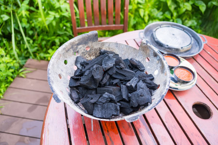 Bowl of black charcoal pieces on a wooden outdoor patio table near a chair with greenery in the backgroundの写真素材
