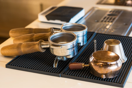 Close-up of espresso portafilters and frothing pitchers on a rubber mat in a coffee shop setting with modern equipmentの写真素材