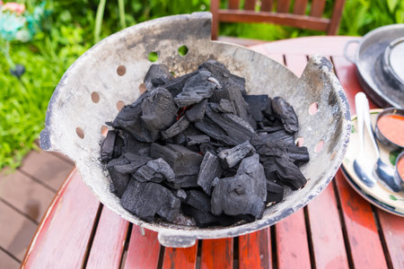 Bowl of black charcoal pieces on a wooden outdoor patio table near a chair with greenery in the backgroundの写真素材